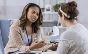 Serious female counselor gestures while talking with Caucasian female client. The counselor is holding eyeglasses and a pen. They are discussing serious issues.